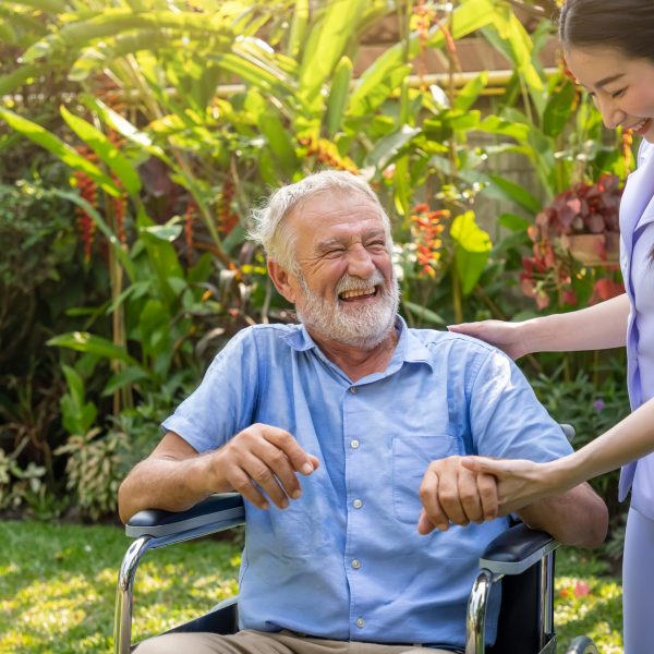 Happy nurse holding laughing elderly man hand on wheelchair in garden at nursing home